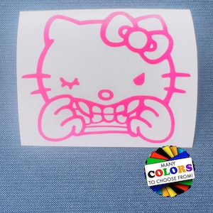 May include: A bright pink vinyl decal featuring a winking Hello Kitty character with a visible set of teeth, positioned above a small circular graphic with the text MANY COLORS TO CHOOSE FROM, displayed on a white rectangular backing sheet.