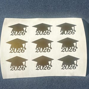 以下が含まれることがあります： 卒業帽子の形の金色のステッカーシートで、それぞれに「2026」の年号が印刷されています。ステッカーは3列3行に配置され、卒業帽のデザインになっています。
