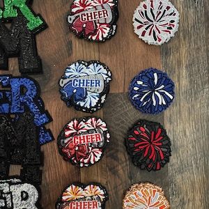 May include: A collection of cheerleading patches and sequined letters. The patches feature the word "CHEER" with pom-pom designs in various colors, including red, white, blue, and orange. The sequined letters spell out "R" and "ER" in green, blue, and black.