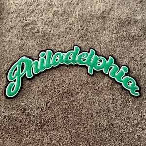 May include: A large, arched embroidered patch featuring the word Philadelphia in a cursive script. The lettering is bright green with a thick white outline and a black border, designed for application onto fabric surfaces.