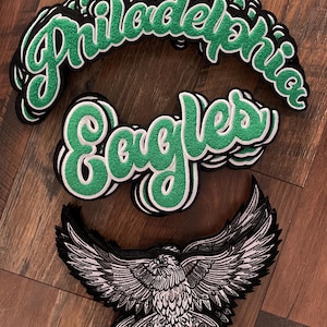 May include: A collection of embroidered patches featuring the words "Philadelphia" and "Eagles" in green script with white and black borders. An eagle patch with outstretched wings is also included, showcasing detailed black and white linework.