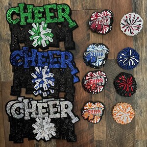 May include: A collection of cheerleading-themed patches and appliqués. The patches feature the word "CHEER" in various colors, including green, blue, and silver, with matching pom-pom designs. The appliqués spell out "MOM" in black.