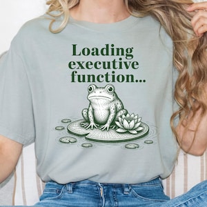 May include: A light sage green short-sleeve t-shirt featuring a dark green graphic of a frog sitting on a lily pad next to a lotus flower. The text "Loading executive function..." is printed in dark green above the frog illustration.