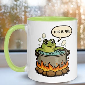 May include: White ceramic mug with a green handle and interior. The mug features a cartoon frog in a pot over a fire, with the text "THIS IS FINE" in a speech bubble. The design is outlined in black.