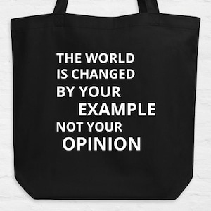 Puede incluir: Bolso tote negro con el texto blanco: "THE WORLD IS CHANGED BY YOUR EXAMPLE NOT YOUR OPINION". El bolso tiene dos asas negras y está hecho de un material resistente.