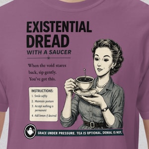 May include: Mauve t-shirt with the text "EXISTENTIAL DREAD WITH A SAUCER" and a vintage illustration of a woman holding a teacup. The shirt includes instructions for handling existential dread and the phrase "GRACE UNDER PRESSURE. TEA IS OPTIONAL. DENIAL IS NOT."