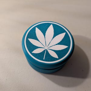 May include: A teal herb grinder with a white cannabis leaf design on the lid. The grinder is round and made of metal. The cannabis leaf is centered on the lid.
