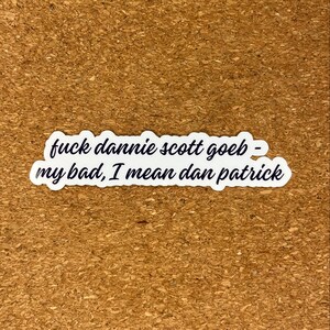 May include: A white sticker with a black, handwritten-style text that reads "fuck dannie scott goeb - my bad, I mean dan patrick". The sticker has a wavy, irregular border and is set against a brown, textured background.
