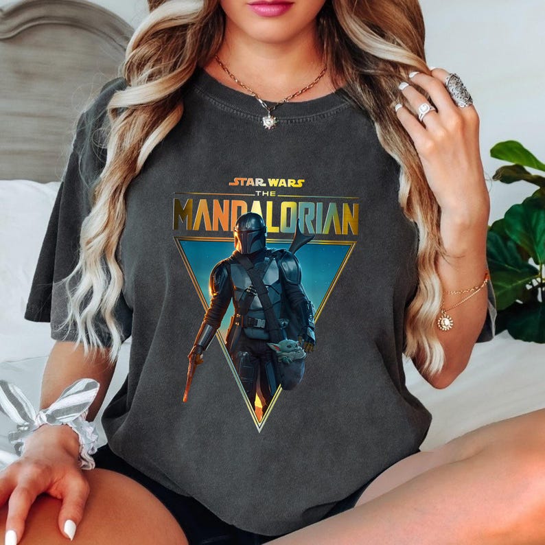 May include: Dark gray t-shirt featuring a graphic from the Star Wars series, "The Mandalorian." The design includes the show's title and an image of the Mandalorian and Grogu within a gold-edged triangle. The shirt is a crew neck.