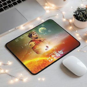 May include: Rectangular mousepad featuring a person in a red and yellow space suit, with the text "BELIEVE IN THE HARLY MAR PROJECT". The background has a planet and a sun-like design.