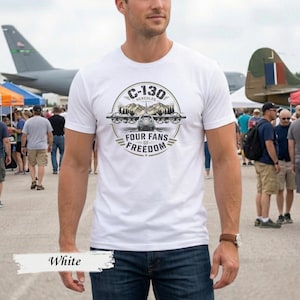Könnte beinhalten: Ein weißes Kurzarm-T-Shirt mit einer kreisförmigen Grafik auf der Brust. Das Design zeigt ein C-130 Hercules Flugzeug mit vier Propellern, Bergen und dem Text C-130 HERCULES FOUR FANS OF FREEDOM in schwarzer und olivgrüner Druckfarbe.