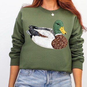 May include: Olive green sweatshirt featuring a detailed appliqu&eacute; of a mallard duck. The duck design incorporates various patterns, including plaid, floral, and striped fabrics, with a yellow beak and a brown floral body. The sweatshirt has long sleeves.