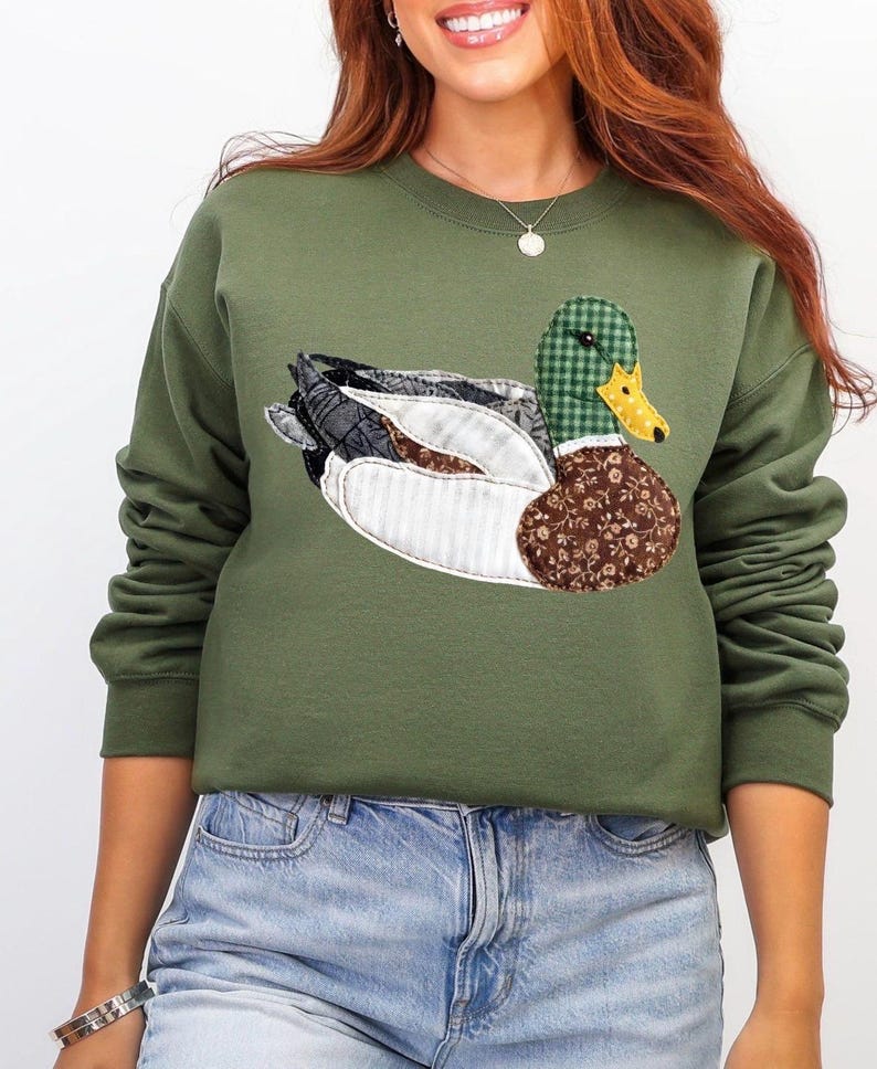 May include: Olive green sweatshirt featuring a detailed appliqu&eacute; of a mallard duck. The duck design incorporates various patterns, including plaid, floral, and striped fabrics, with a yellow beak and a brown floral body. The sweatshirt has long sleeves.