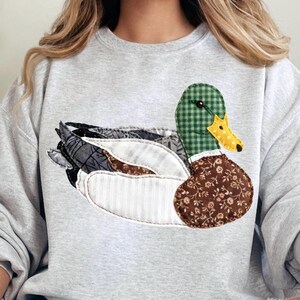 May include: A light gray sweatshirt featuring a patchwork duck design. The duck has a green checkered head, a yellow and white dotted beak, and a brown floral patterned chest. The wings are made of various fabric patterns.
