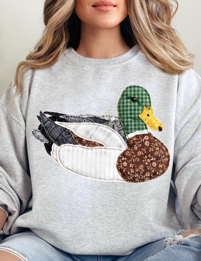 May include: A light gray sweatshirt featuring a patchwork duck design. The duck has a green checkered head, a yellow and white dotted beak, and a brown floral patterned chest. The wings are made of various fabric patterns.
