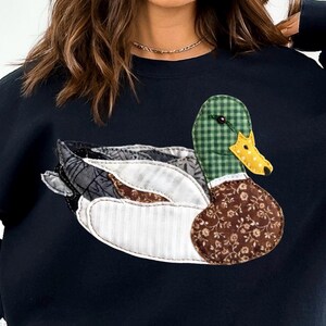 May include: Black sweatshirt featuring a patchwork duck design. The duck has a green checkered head, a yellow beak, and a brown floral patterned body. The wings are a mix of patterned fabrics, including white stripes and gray patterns.