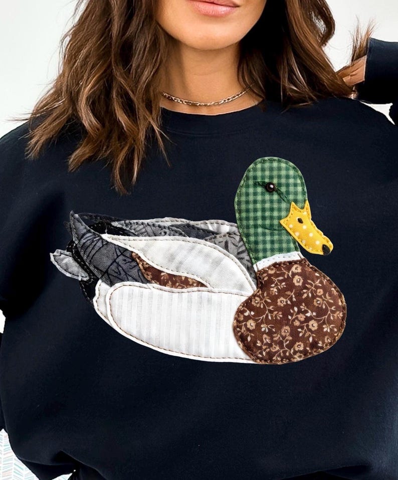 May include: Black sweatshirt featuring a patchwork duck design. The duck has a green checkered head, a yellow beak, and a brown floral patterned body. The wings are a mix of patterned fabrics, including white stripes and gray patterns.