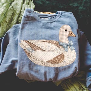 May include: A pale blue sweatshirt with a decorative duck applique. The duck has a checkered head, a brown beak, and a light blue bow. The duck's body is made of various patterned fabrics, including polka dots and floral prints.