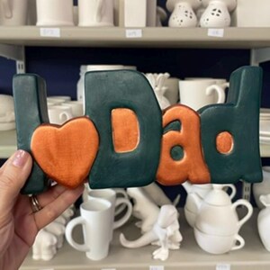 May include: A decorative ceramic sign featuring the text "I heart Dad" in a bold, block-letter style. The letters are painted in a deep forest green, while the heart symbol and the centre of the letters are finished in a metallic copper paint.
