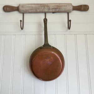 May include: A round copper pan with a long, dark metal handle hanging from a hook. Above the pan, a wooden rolling pin with dark brown handles is mounted horizontally on a wall rack featuring three metal hooks against a white panelled wall.
