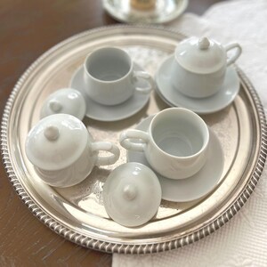 Peut inclure: Un plateau argenté présente de petites tasses à thé blanches, des soucoupes et des pots couverts. Les tasses ont des anses incurvées. Les couvercles des pots ont de petits boutons arrondis. L'ensemble suggère un service à thé.