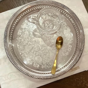 Peut inclure: Un plateau rond en métal argenté avec un bord torsadé décoratif et un motif floral en relief sur la base. Un pot à lait en verre transparent avec un motif en étoile et une petite cuillère en métal doré sont posés sur le plateau.