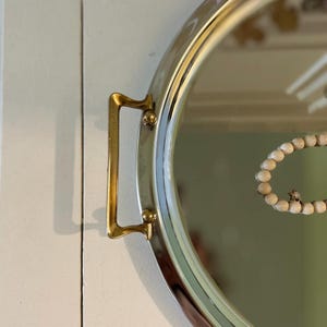 May include: A circular mirror with a silver frame and a gold-coloured handle. A beaded bracelet with a small charm is reflected in the mirror. The mirror is mounted on a white surface.