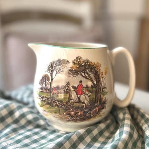 May include: Cream-colored ceramic pitcher with a hunting scene illustration. The pitcher features a handle and a green rim. The illustration depicts a rider on a horse, hounds, and a landscape with trees and a fence.