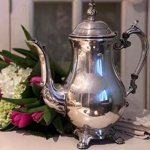 May include: A polished silver coffee pot with ornate detailing, including a curved spout and handle. The pot is displayed next to a bouquet of pink tulips and white flowers, creating a classic and elegant aesthetic.