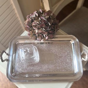 May include: A rectangular silver-toned metal serving tray with ornate embossed floral patterns and two side handles. A small, clear glass square container with a matching lid sits on the left side of the tray surface, reflecting light.