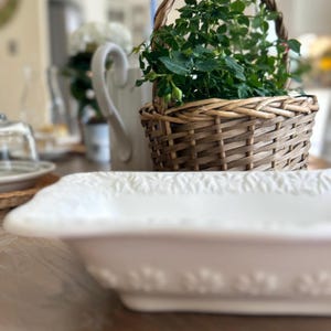 May include: A white rectangular serving dish with a floral pattern on the rim. A woven basket with a handle, filled with green plants, is in the background. A white jug and other tableware are also visible.
