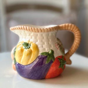 May include: A ceramic jug with a woven basket design, decorated with a yellow pepper, purple aubergine, and red tomato. The handle and rim have a woven texture. A decorative kitchenware item.