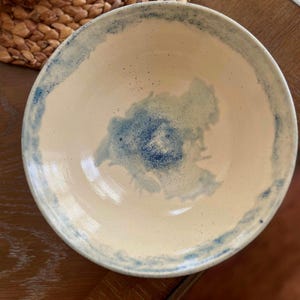 May include: A handmade ceramic bowl with a cream interior and a blue watercolour design. The rim and centre of the bowl feature a blue wash. The bowl is round and appears to be a decorative piece.