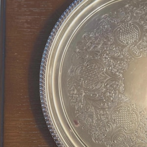 Peut inclure: Un plateau de service rond en métal argenté présentant un motif floral et des volutes en relief complexe le long du bord. Le contour du plateau est fini avec un rebord décoratif de style corde, offrant un contraste texturé au centre lisse.