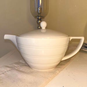 May include: A cream-coloured ceramic teapot featuring a rounded body with three horizontal embossed lines, a matching lid with a small spherical knob, a short spout, and a flat-topped handle extending from the side of the vessel.
