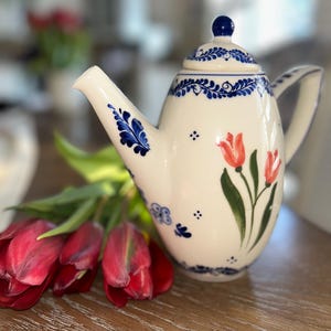 May include: Cream ceramic teapot with blue floral and tulip patterns. The teapot features a curved spout, a handle, and a lid with a blue knob. Red tulips with green stems and leaves are next to the teapot.