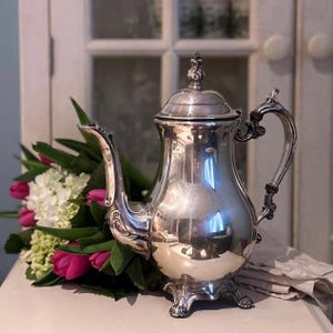 May include: A polished silver coffee pot with decorative features, including a curved handle and spout. The pot is presented with a bouquet of pink tulips and white flowers, creating a classic and elegant look.