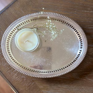Peut inclure: Un plateau ovale en métal argenté avec un bord ajouré décoratif et un motif floral gravé sur la surface. Un petit pot en verre contenant une bougie couleur crème et une branche de fleurs séchées gypsophile reposent sur le plateau.