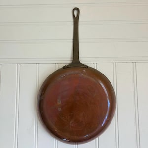 Peut inclure: Une poêle ronde en cuivre avec une patine sombre et usée, dotée d'un long manche en métal foncé. La surface de la poêle présente des traces d'utilisation avec des variations subtiles de couleur et de texture, incluant une forme circulaire.