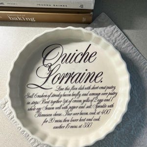 May include: A white ceramic quiche dish with a fluted edge, inscribed with "Quiche Lorraine" and a recipe. The dish is on a white cloth. Recipe measurements are in imperial units. Books are visible in the background.