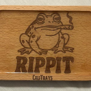 Pode incluir: Bandeja retangular de madeira com cantos arredondados. A bandeja apresenta um sapo fumando um cigarro, com a palavra "RIPIT" e "CALITRAYS" gravadas na madeira.