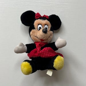 May include: A plush Minnie Mouse doll with black ears, a red bow, and a red and white polka dot dress. The doll has a tan face, white hands, and yellow feet. A small tag is attached to the doll.