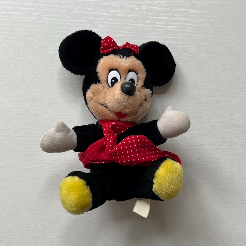 May include: A plush Minnie Mouse doll with black ears, a red bow, and a red and white polka dot dress. The doll has a tan face, white hands, and yellow feet. A small tag is attached to the doll.