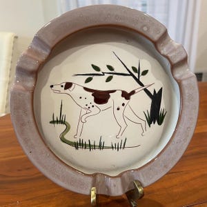 Peut inclure: Cendrier rond en céramique avec un bord violet clair et une illustration centrale d'un chien. Le chien est blanc avec des taches marron, dans un champ avec de l'herbe verte et un arbre. Le cendrier a un support en métal doré.