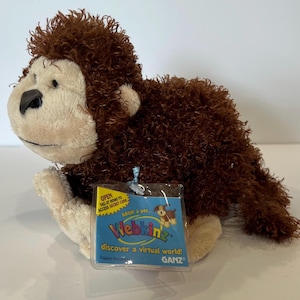 Op de afbeelding: Een bruin en crèmekleurig pluche apenknuffel met krullende vacht, een glad gezicht en een zwarte neus. Aan de voorzijde is een doorzichtig plastic label bevestigd met de tekst Webkinz, Adopt a pet, discover a virtual world en GANZ in blauw en geel.