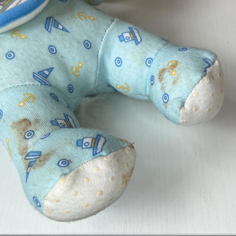 May include: Close-up of a light blue plush toy's leg. The fabric is patterned with small sailboats, anchors, and circles. The foot is white with small yellow dots. There are brown stains on the fabric.