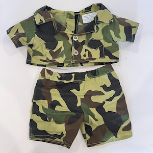 May include: A camouflage print shirt and shorts set featuring a green, brown, and black pattern. The short-sleeved shirt has a collar, two front pockets with flaps, and two silver-toned buttons. A white tag inside the shirt reads Build-A-Bear Workshop.