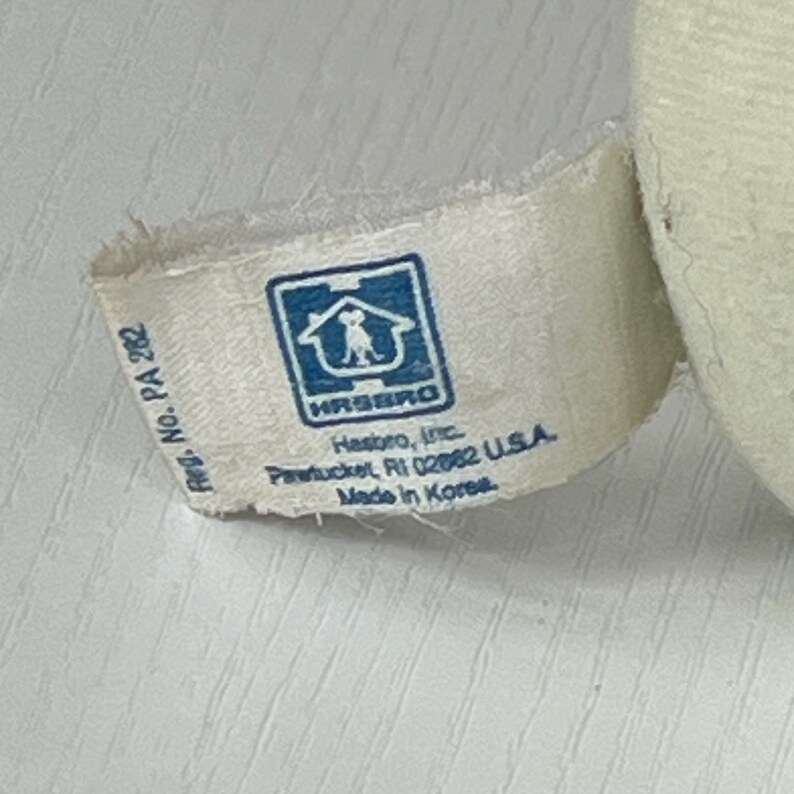 May include: Close-up of a white fabric tag with the Hasbro logo, a blue square with a house and dog silhouette. The tag includes the text "Hasbro, Inc. Pawtucket, RI 02862 U.S.A. Made in Korea."