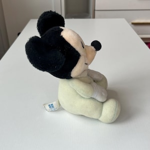 May include: A plush Mickey Mouse toy in a seated position. The toy features black ears, a black nose, and a cream-colored face and body. The toy has a small tag attached to its side.