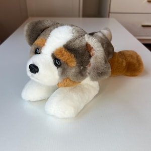 May include: A plush dog toy in a lying position. The toy has a white face with black eyes and a black nose. The ears and top of the head are gray and brown, with orange accents. The paws and body are white, and the tail is brown.